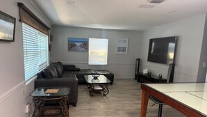 A living room with a sectional sofa, a glass coffee table, a wooden dining table, and a flat-screen TV.