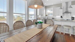Una cocina moderna con armarios blancos, una mesa de comedor de madera con un camino tejido, sillas de mimbre y una lámpara colgante.