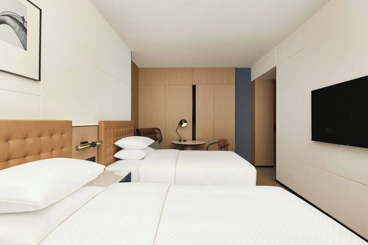 Hotellihuone, jossa on kaksi sänkyä, työpöytä, tuoli, televisio ja seinällä oleva kehystetty kuva.