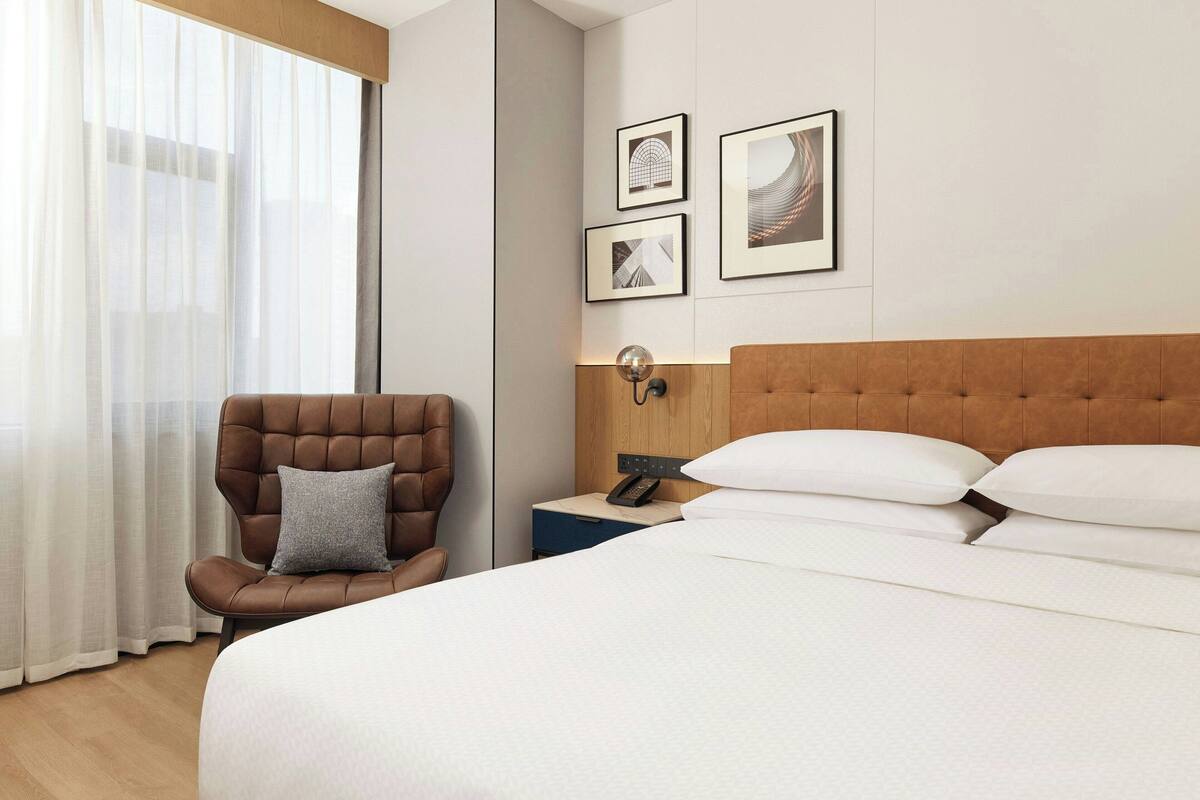 Hotellihuone, jossa on suuri sänky, puinen pääty, yöpöytä lampun kanssa, tyynyillä varustettu tuoli ja ikkuna, jossa on ohuet verhot.