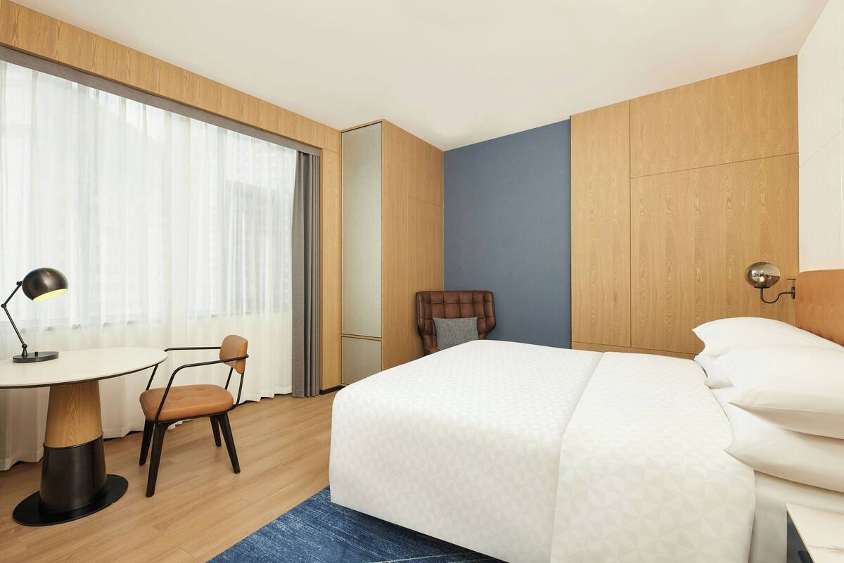 Hotellihuone, jossa on sänky, työpöytä tuolilla sekä suuri ikkuna, jossa on verhot.