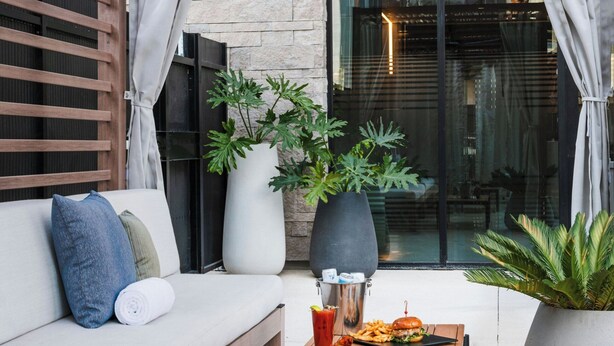 An outdoor seating area with a wooden bench, a table with food, potted plants, and a view of the building's interior through a glass door. - AC Hotel by Marriott Las Vegas Symphony Park (Las Vegas)
