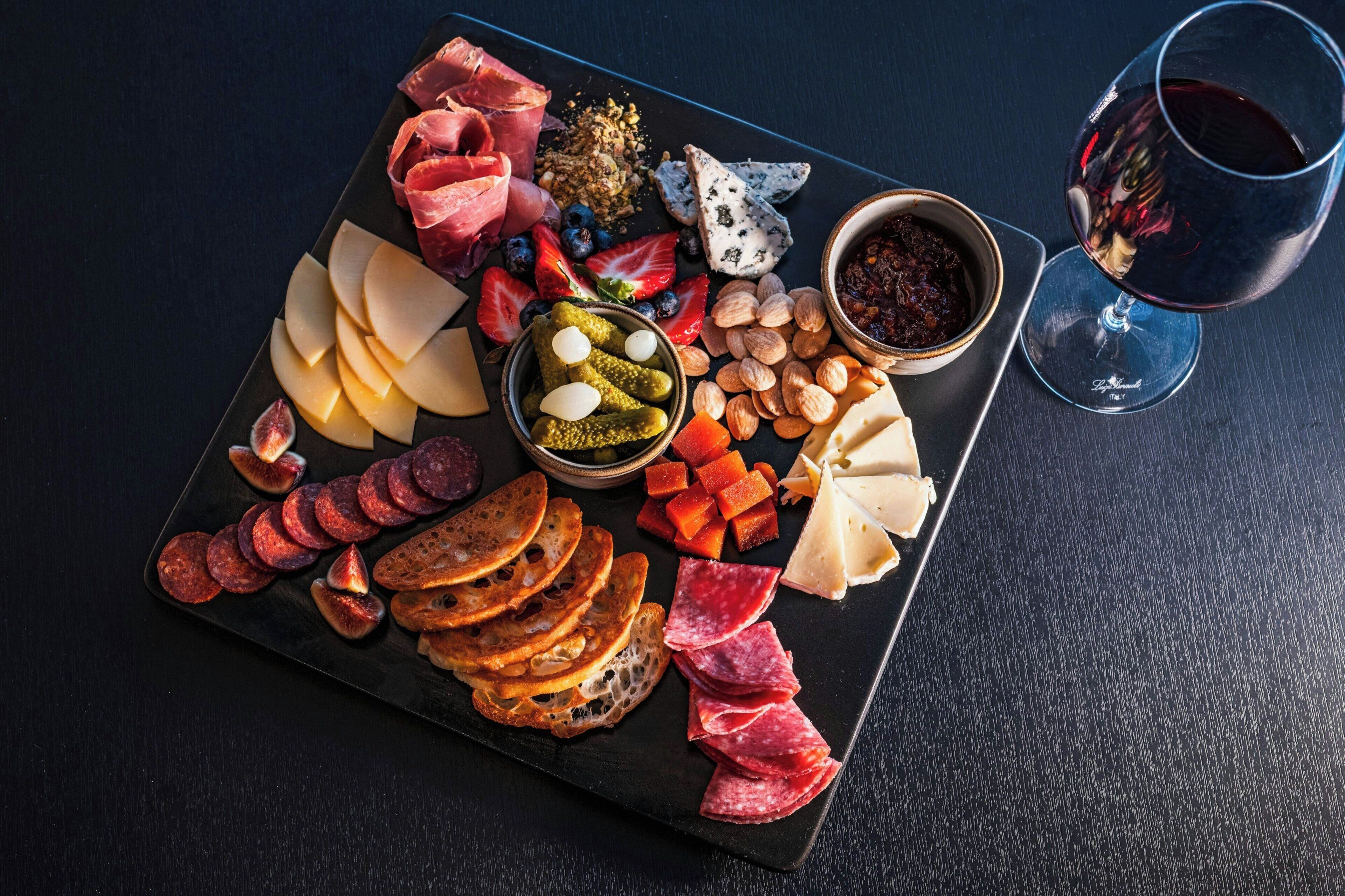 Eine Platte mit verschiedenen Wurstwaren, Käsesorten, Früchten und Nüssen, dazu ein Glas Rotwein.