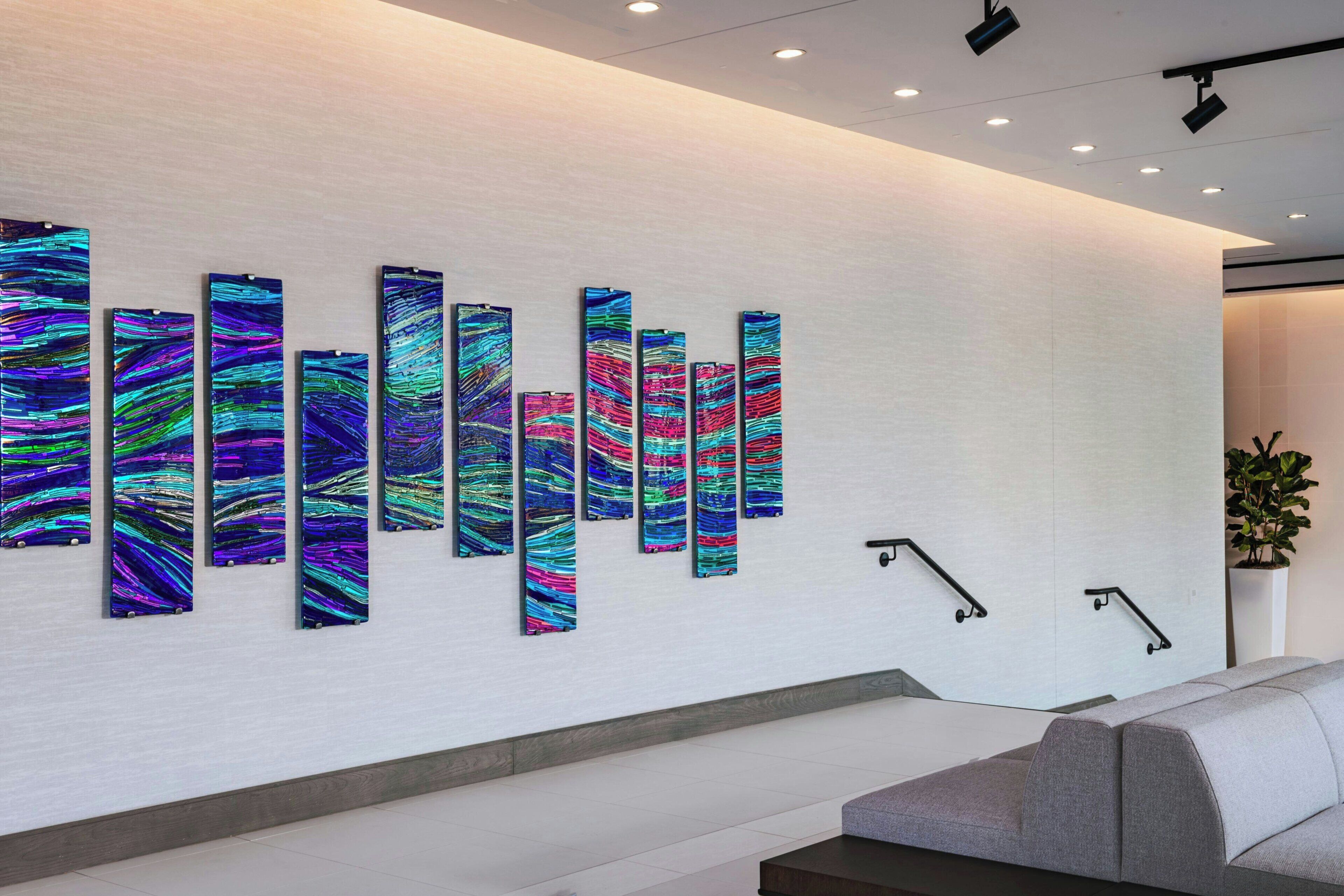 Een modern interieur met een witte muur, een rij kleurrijke abstracte kunstwerken, een grijze bank en een plant.