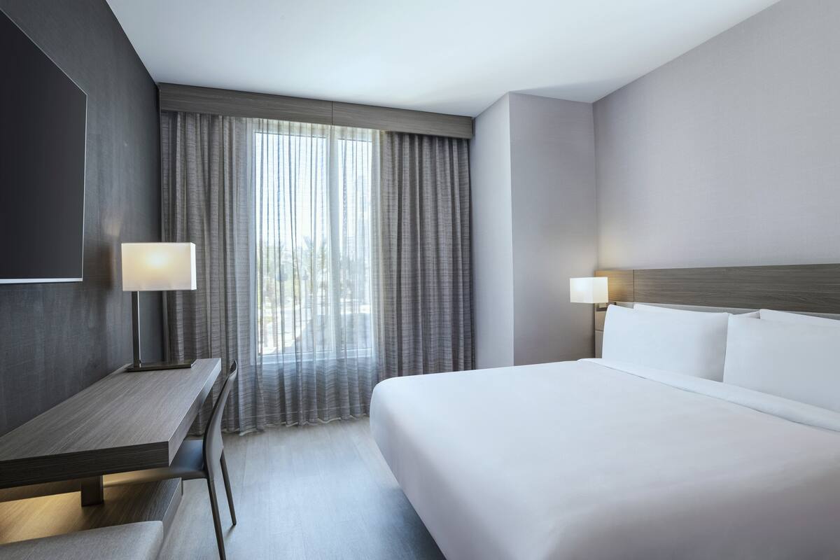 Una camera d'hotel moderna con un letto grande, una scrivania e vista sulla città attraverso tende leggere.