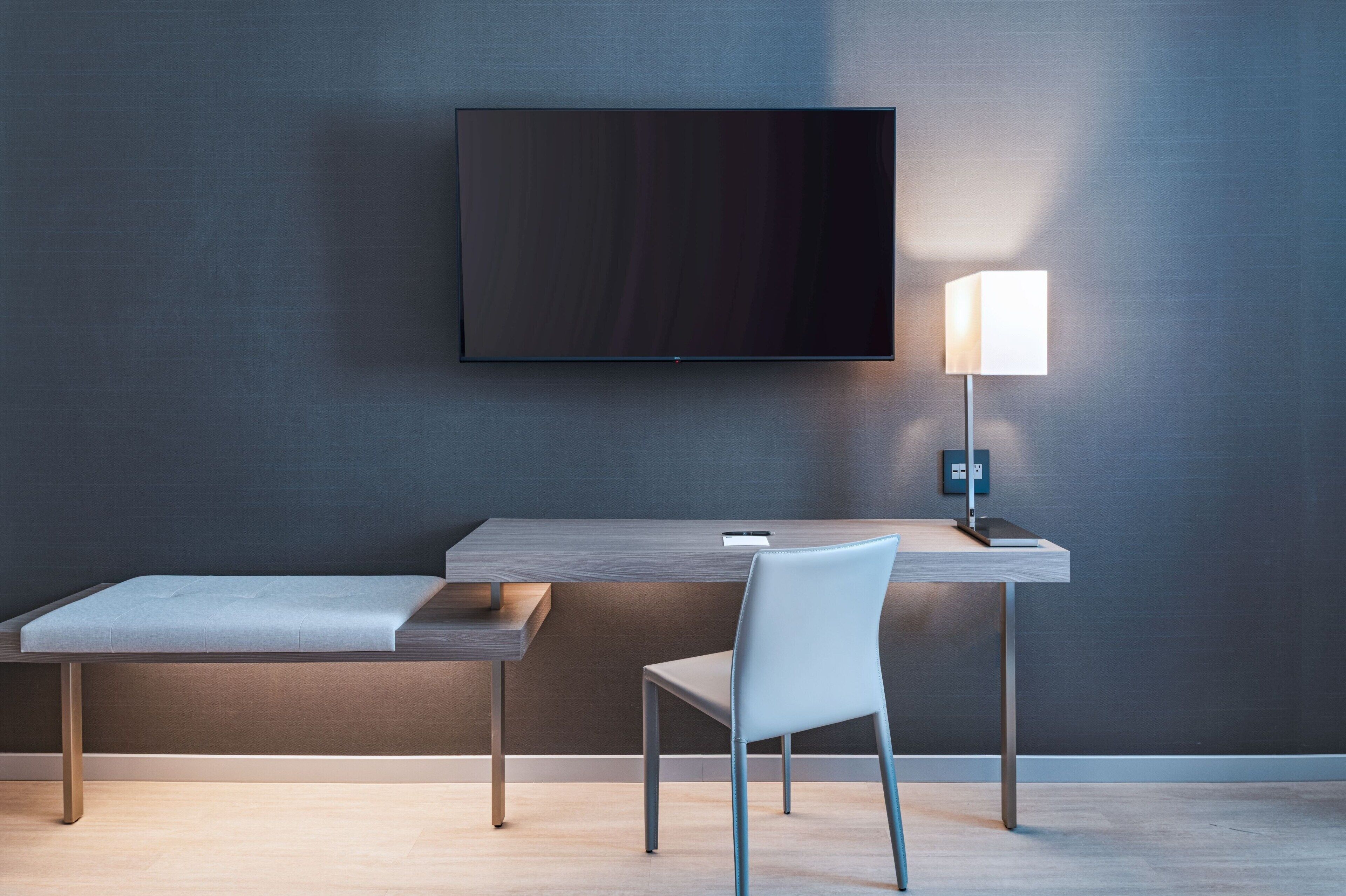 Et moderne værelse med et vægmonteret fladskærms-tv, et skrivebord med bænk, en stol og en gulvlampe.