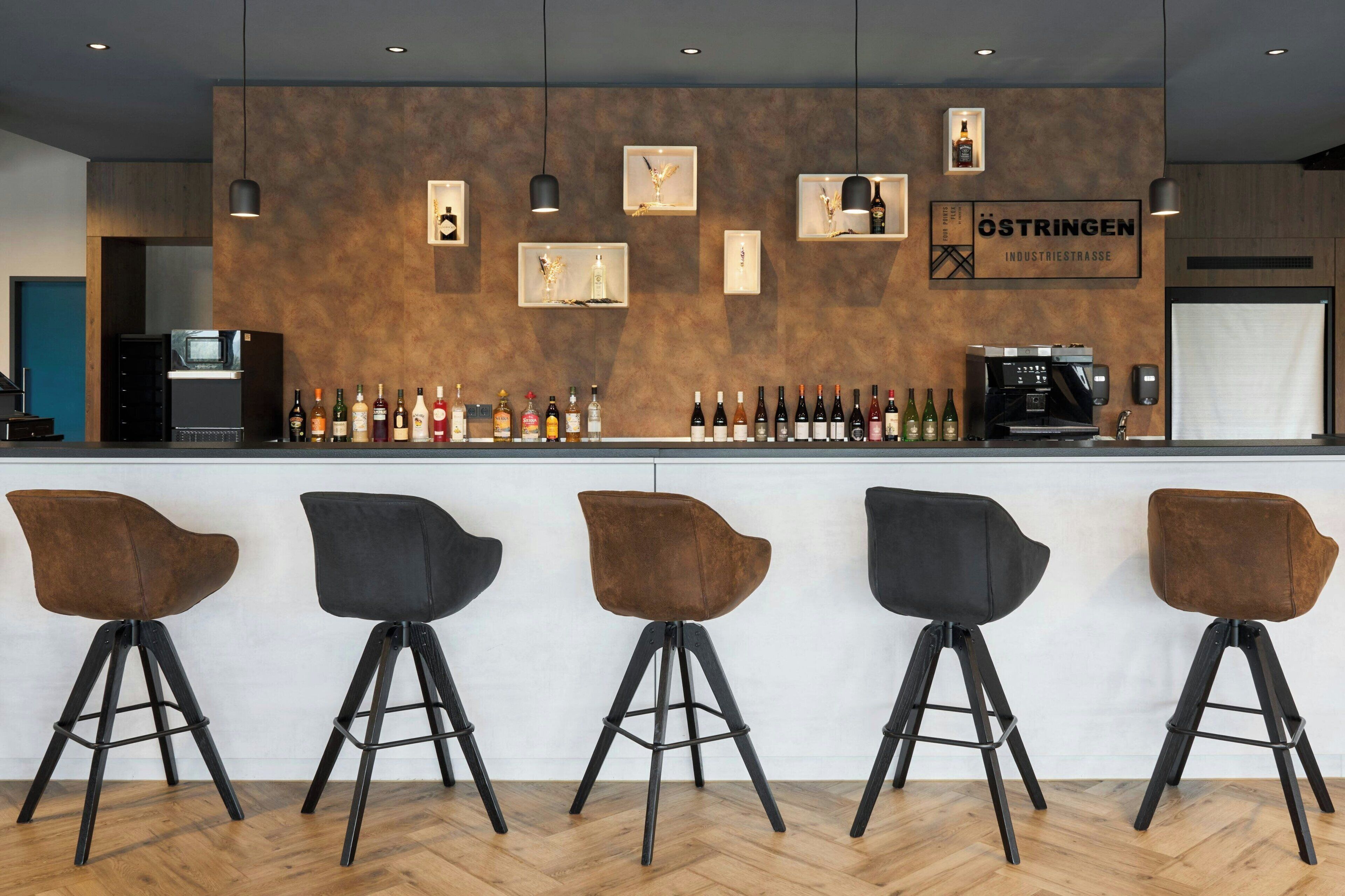 Eine Bar mit vier Hockern, einer Theke mit Flaschen und einem Schild mit der Aufschrift 'ÖSTRINGEN INDUSTRIESTRASSE'.