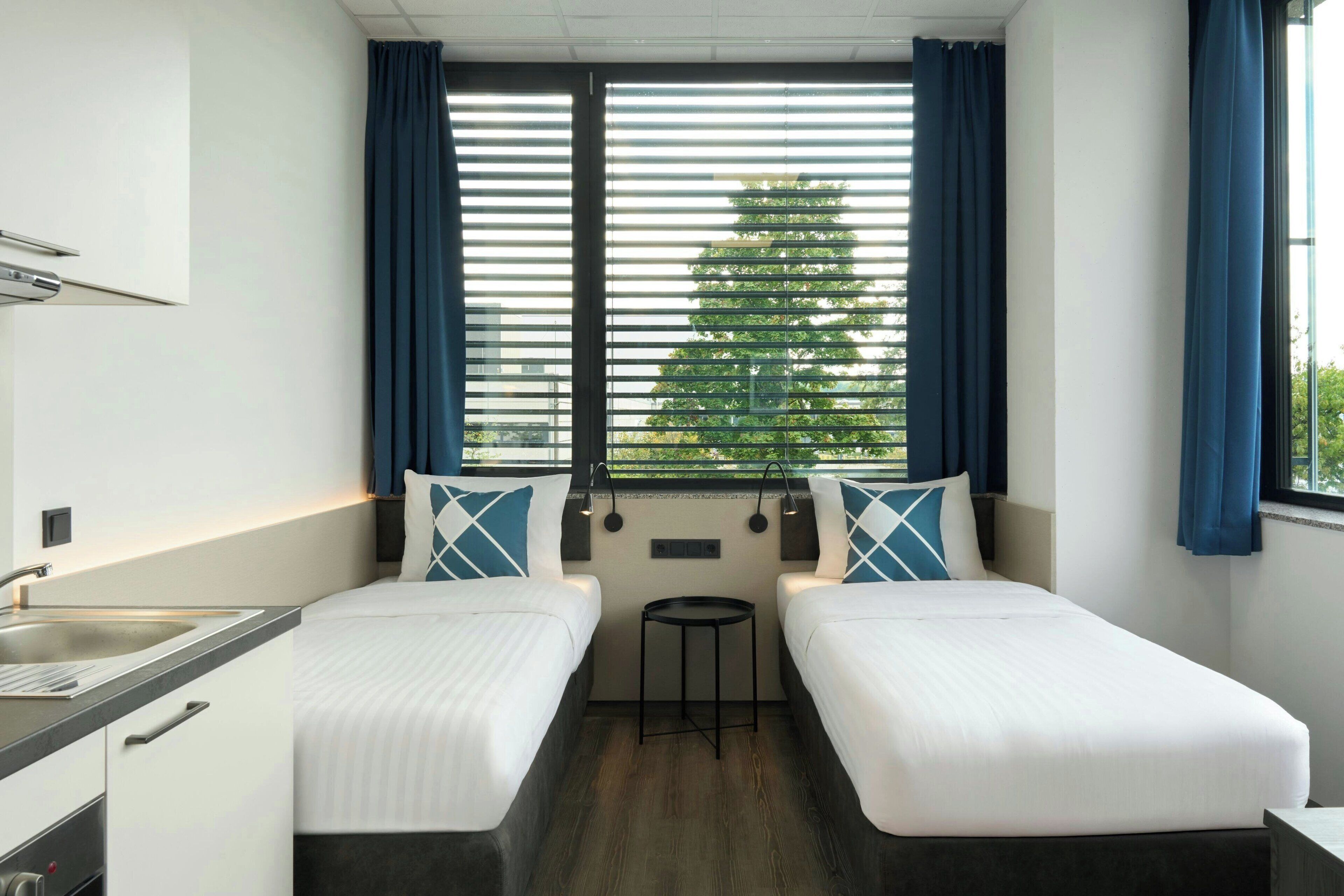 A hotel room with two single beds, a small round table, a window with blinds, and a kitchenette with a sink and cabinets.