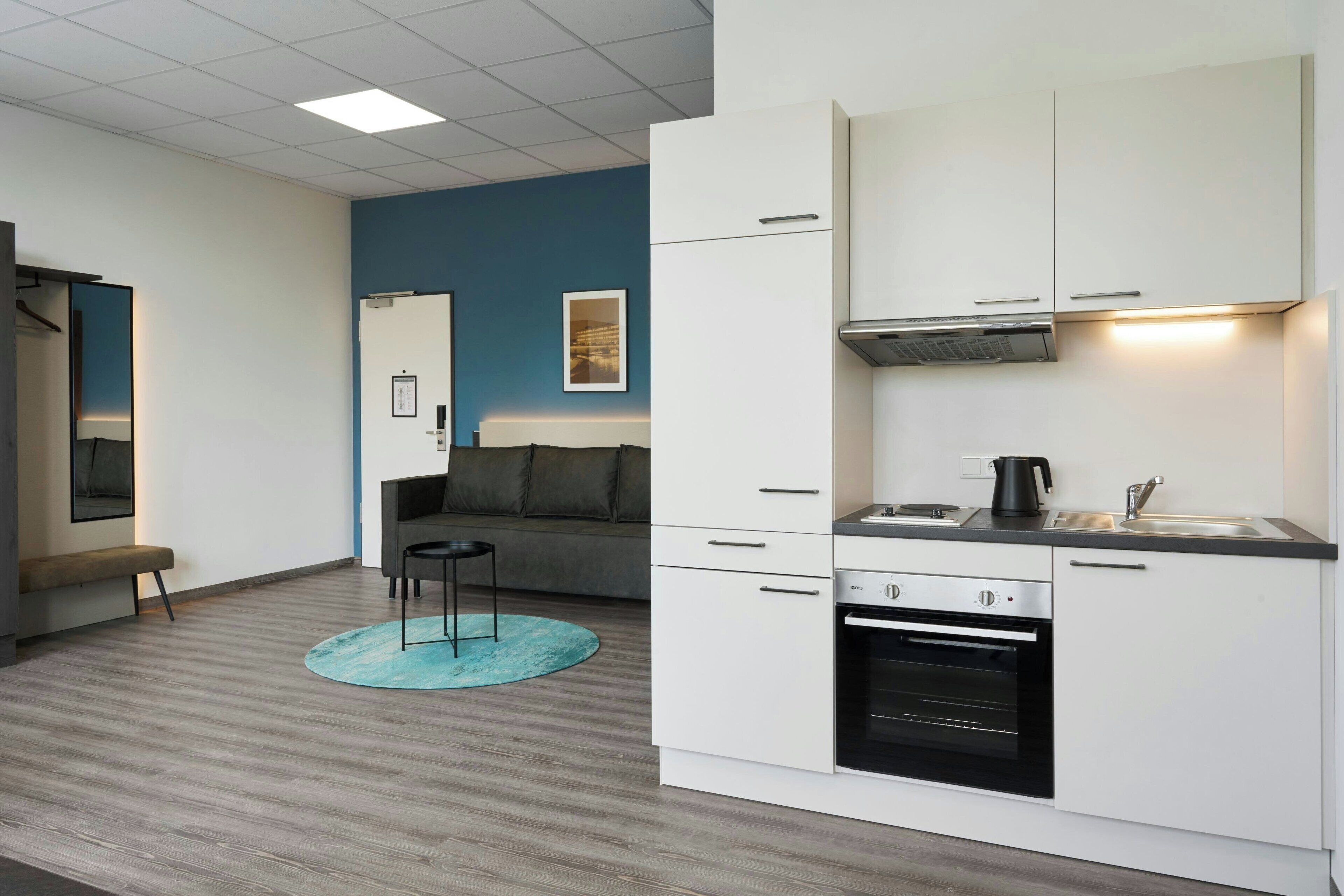 A modern kitchen with white cabinets, a built-in oven, and a coffee maker. There is a blue rug on the floor and a gray sofa in the background.