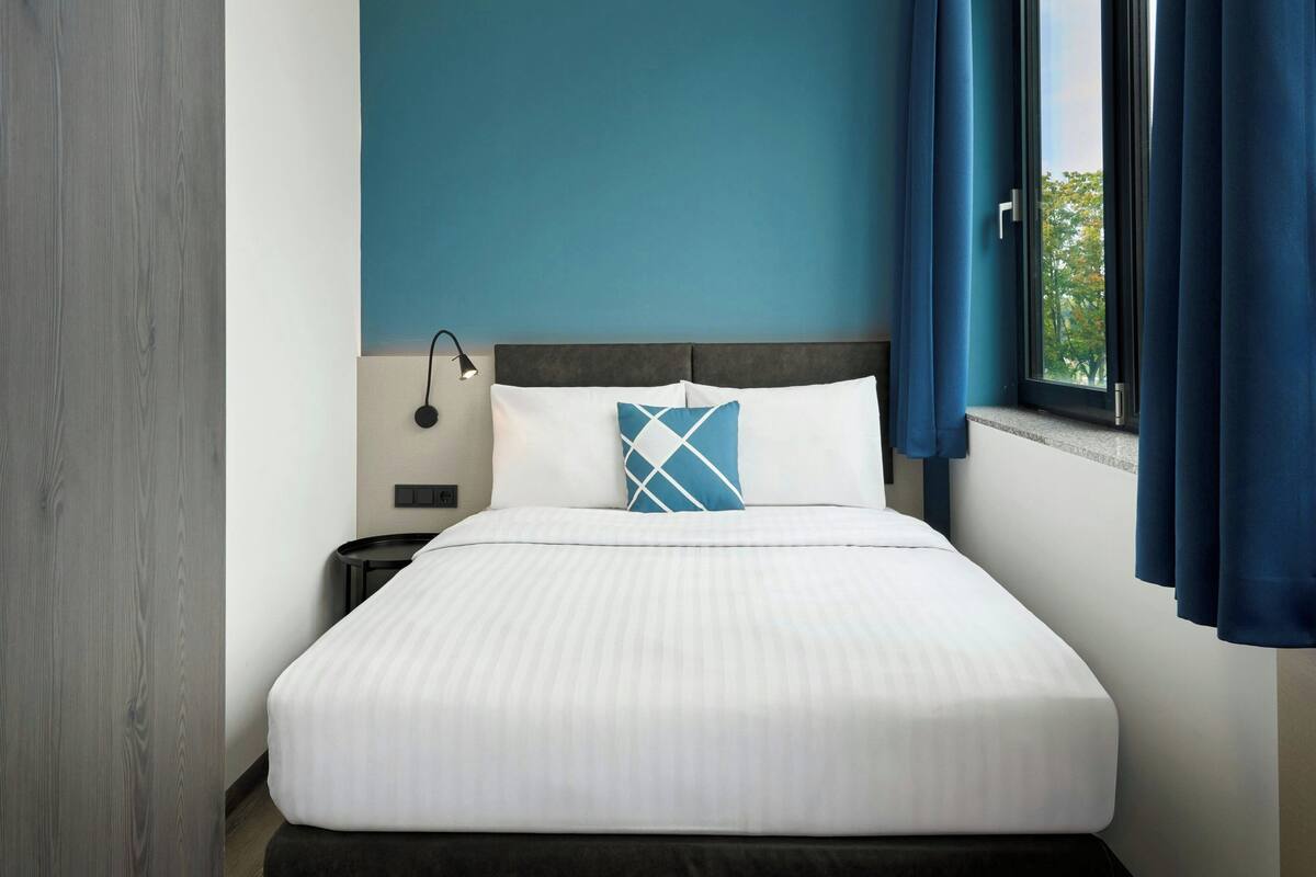Ein ordentlich bezogenes Bett mit weißer Bettwäsche und einem blauen geometrischen Kopfkissen, eine Nachttischlampe und ein Fenster mit blauen Vorhängen.