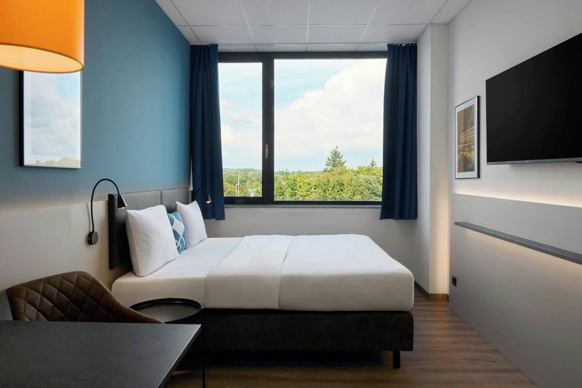 Ein Hotelzimmer mit Bett, Schreibtisch, Stuhl, Fernseher und Fenster mit Blick ins Grüne.