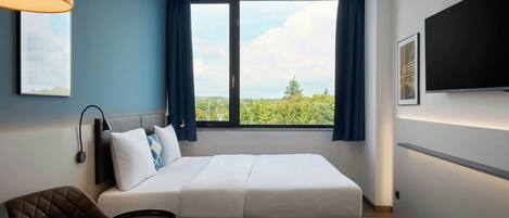 Une chambre d’hôtel équipée d’un lit, d’un bureau, d’une chaise, d’une télévision et d’une fenêtre donnant sur un paysage verdoyant.