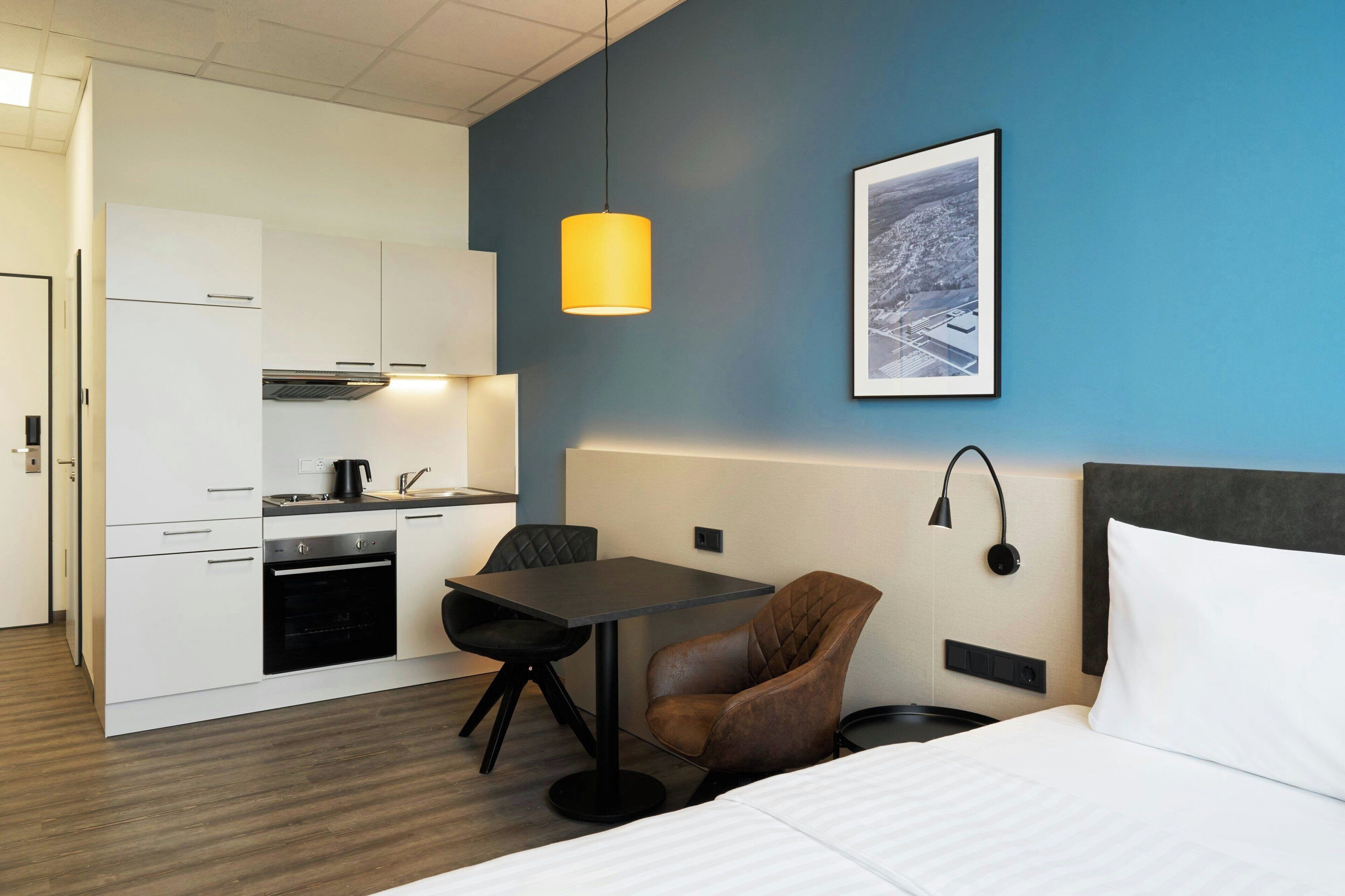 A compact hotel room with a kitchenette, a dining table, a single brown chair, a bed with white bedding, a blue accent wall, and a framed picture.