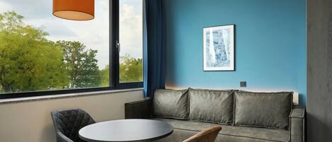 A modern room with a grey sofa, a round table, and a chair. There is a window with a view of trees and a blue wall.