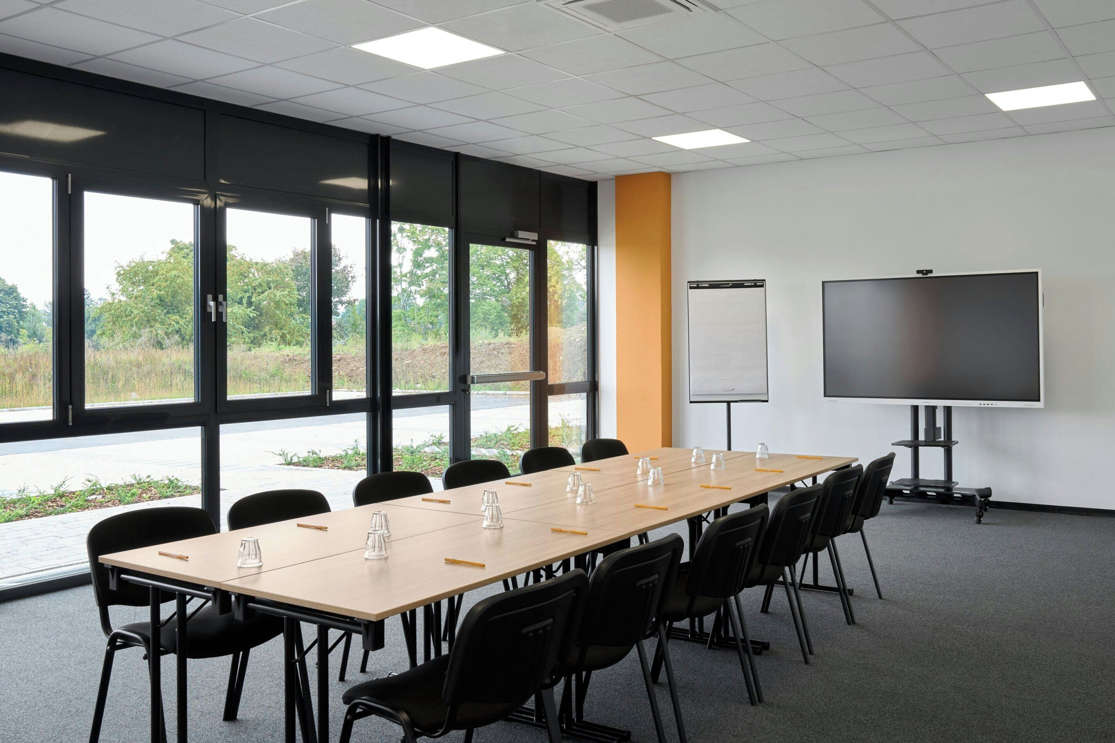A conference room with a long table, chairs, a projector screen, and large windows offering a view of greenery.