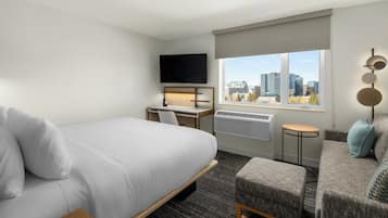 A hotel room with a bed, a desk with a chair, a sofa, a TV mounted on the wall, and a window with a city view.