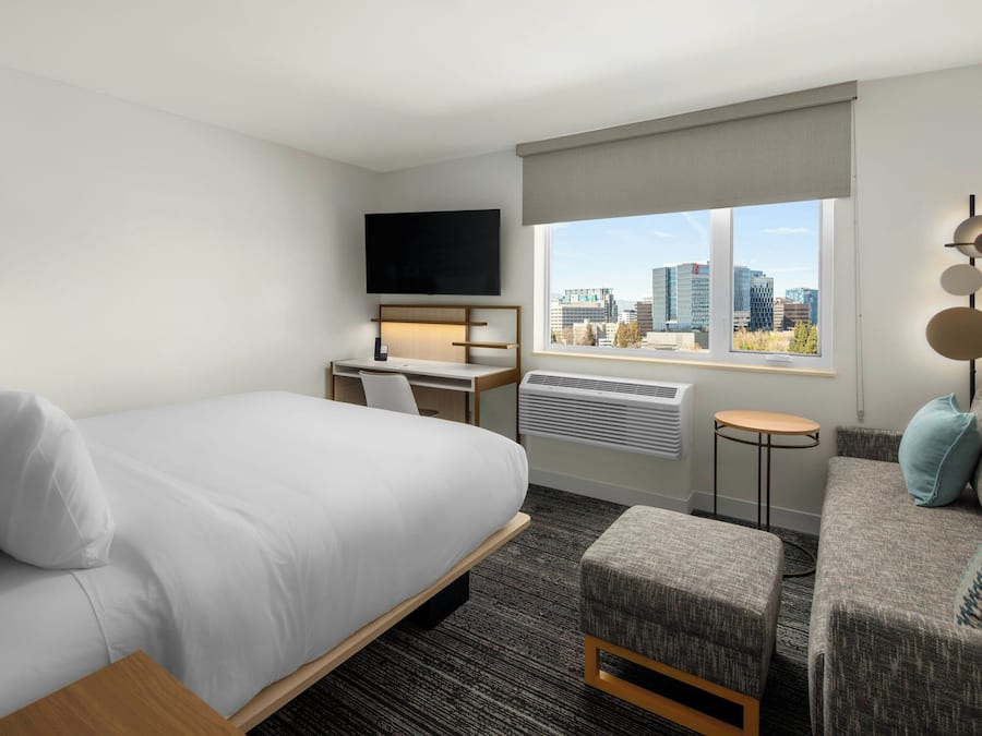 A hotel room with a bed, a desk with a chair, a sofa, a TV mounted on the wall, and a window with a city view.