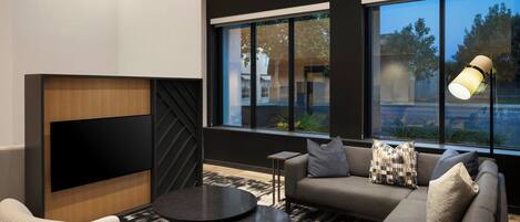 Un salon moderne avec un canapé, un fauteuil et une table basse, de grandes fenêtres et un téléviseur à écran plat fixé au mur.