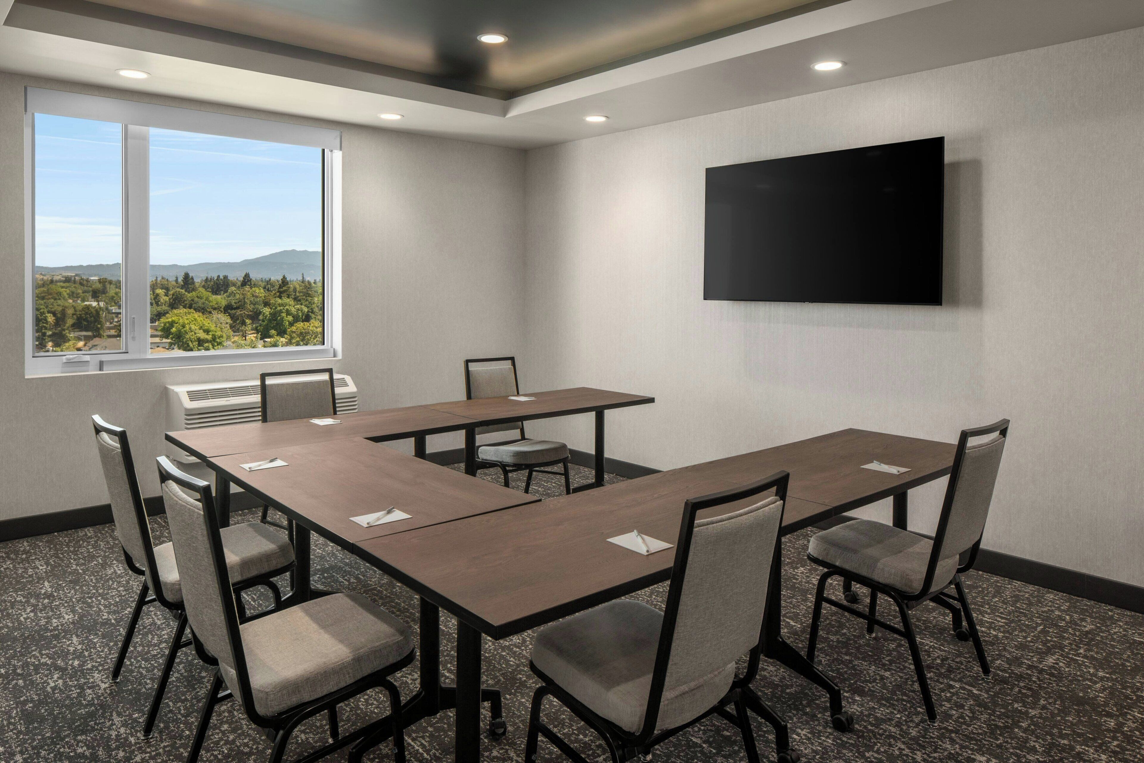 A conference room with a large table, chairs, a flat-screen TV, and a window with a view of trees and mountains.