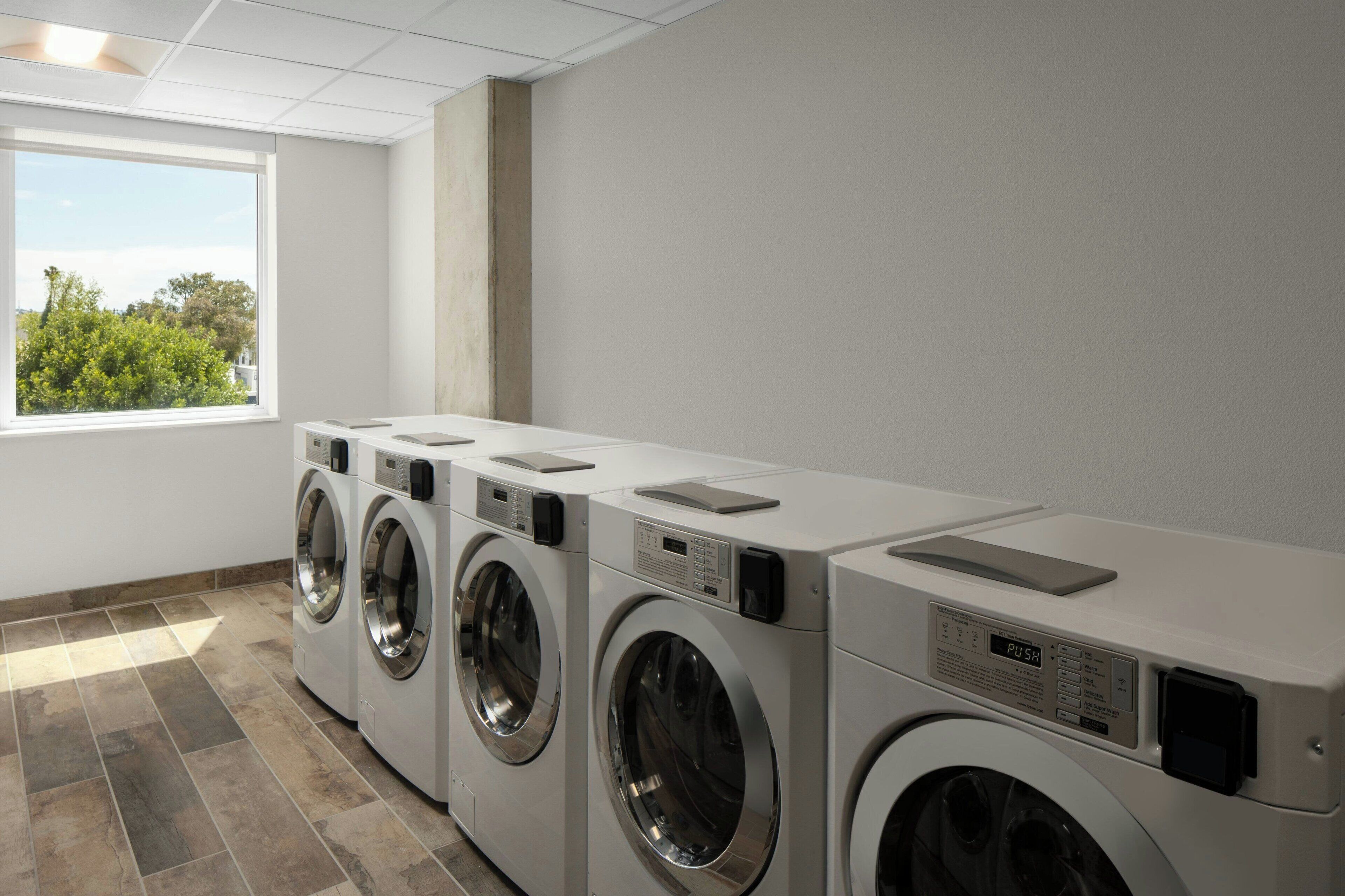 A row of front-loading washing machines in a laundry room with a large window and a view of trees.