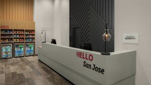 Un mostrador de recepción con un cartel que dice 'HELLO San Jose', un minibar con aperitivos y bebidas, y un cartel en la pared que indica 'MARRIOTT BONVOY'.