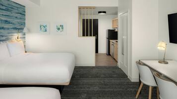 A hotel room with a large bed, a desk with two chairs, a kitchenette, and artwork on the walls.