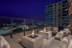 A rooftop terrace with modern seating, two square fire pits, and a city view at night.