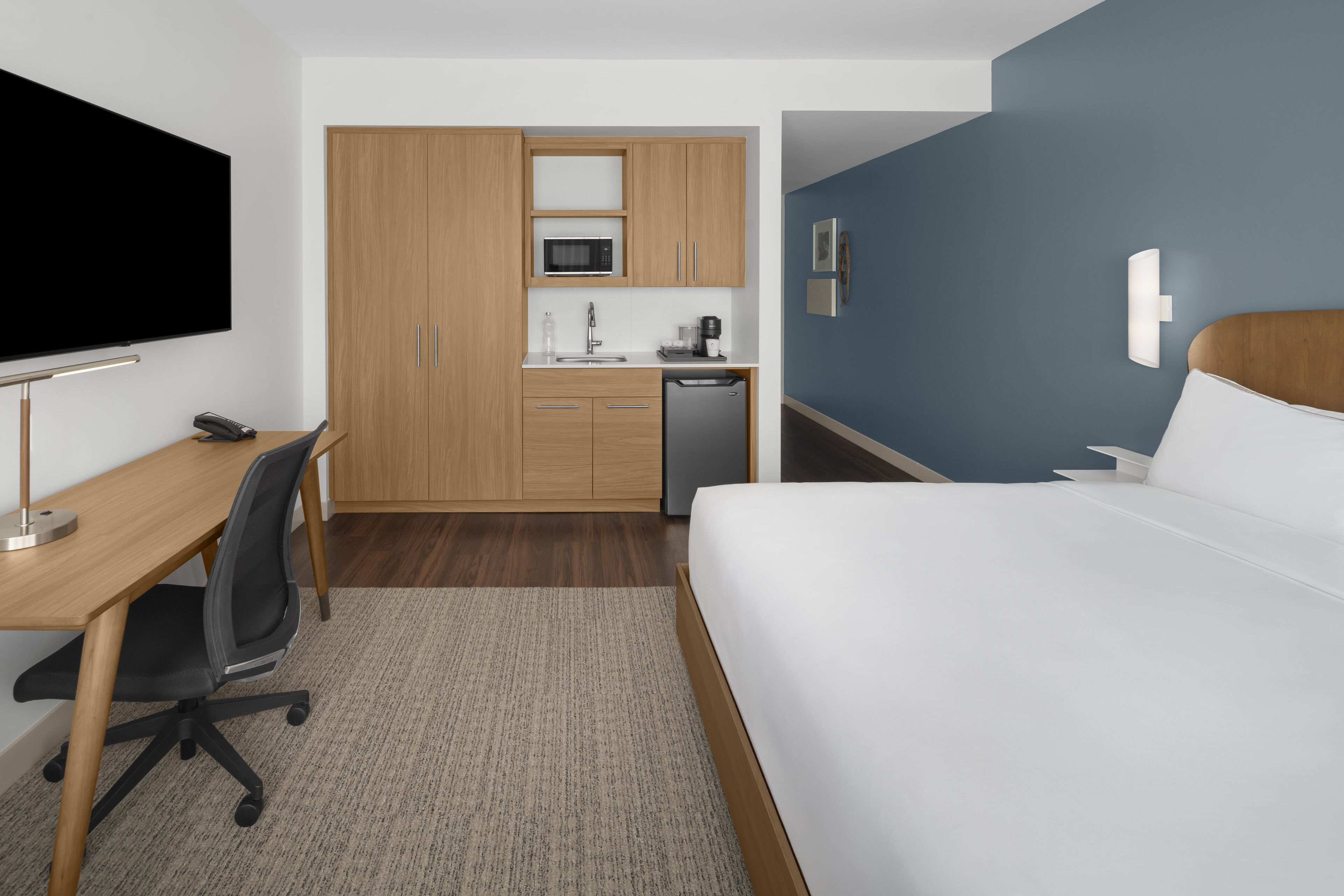 A hotel room with a bed, a desk, a chair, a TV, a microwave, a sink, and a small kitchenette.