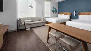 A modern hotel room with a wooden desk, two beds, a sofa, and a large window with a view.