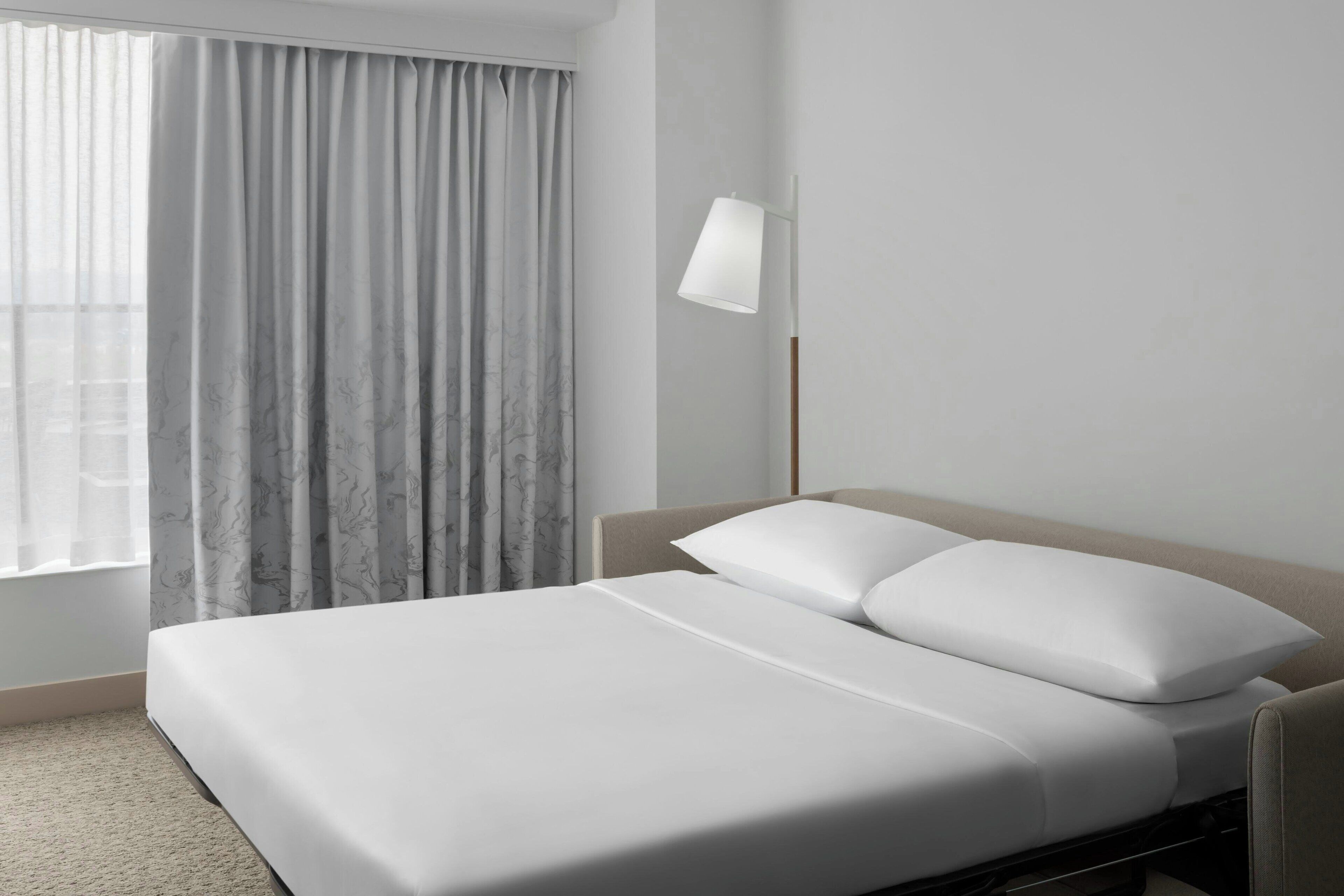 A modern hotel room with a neatly made bed, a bedside lamp, and a view of the city through sheer curtains.
