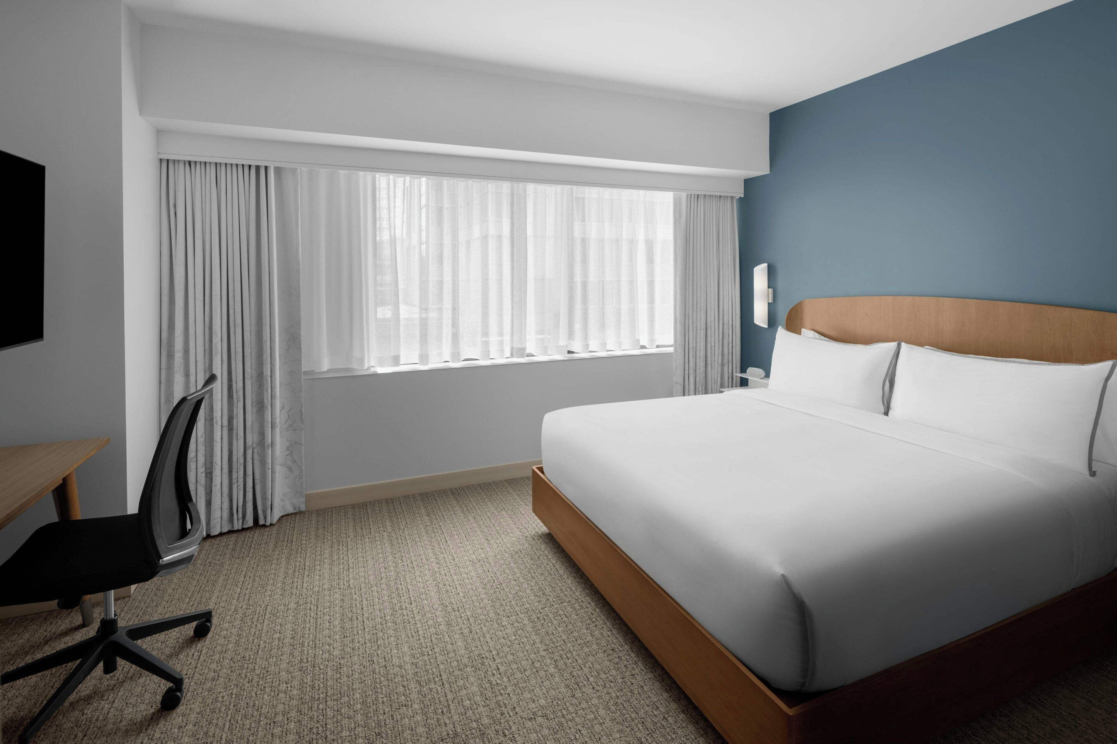 Habitación de hotel con una cama grande, un escritorio, una silla y un ventanal con cortinas.