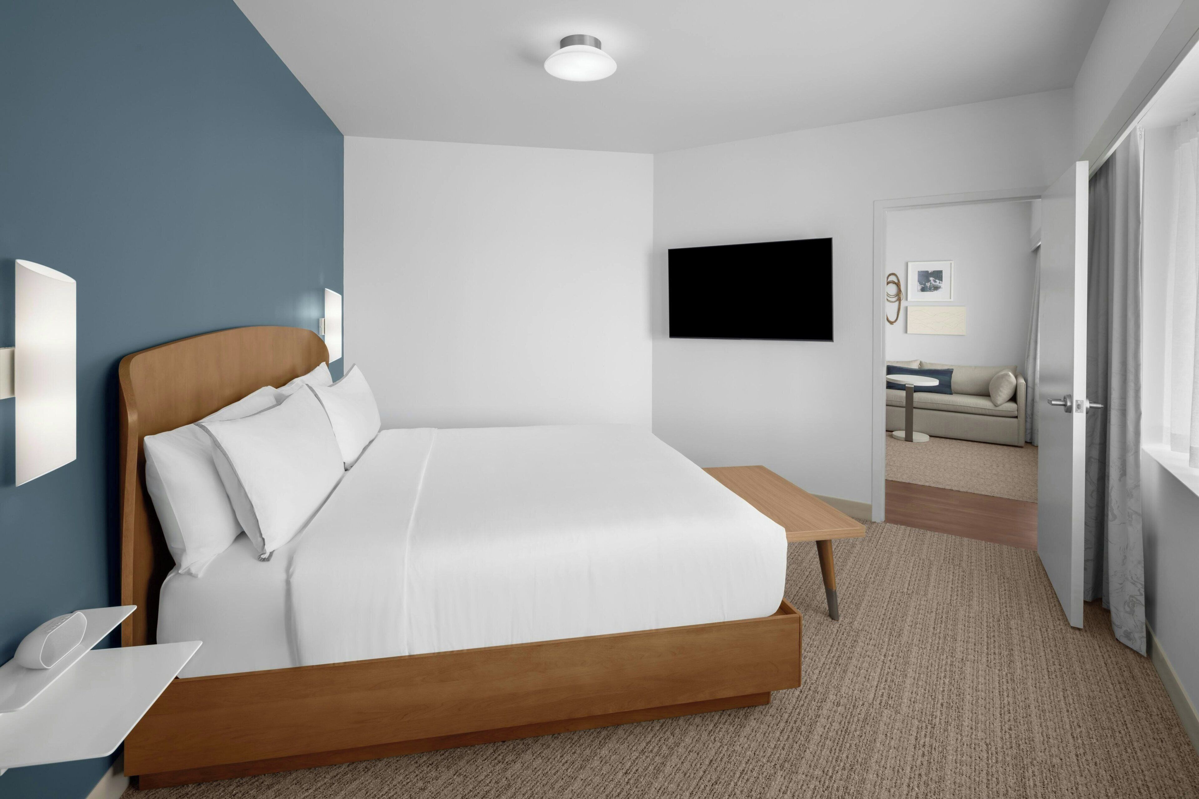 Habitación de hotel moderna con cama de madera, televisor de pantalla plana, mesita de noche y vista al área de estar contigua.