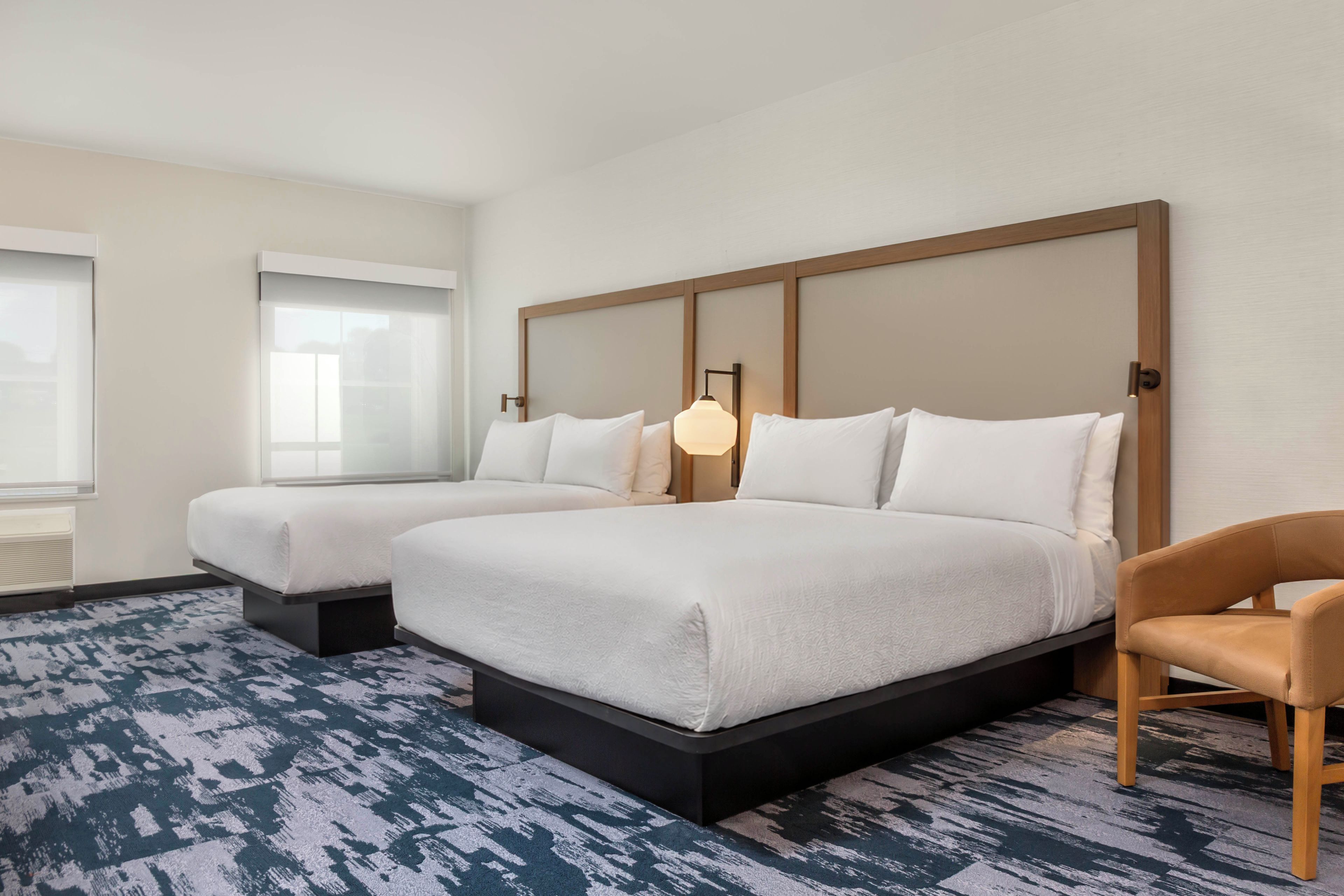 A hotel room with two beds, a chair, a window, and a lamp.
