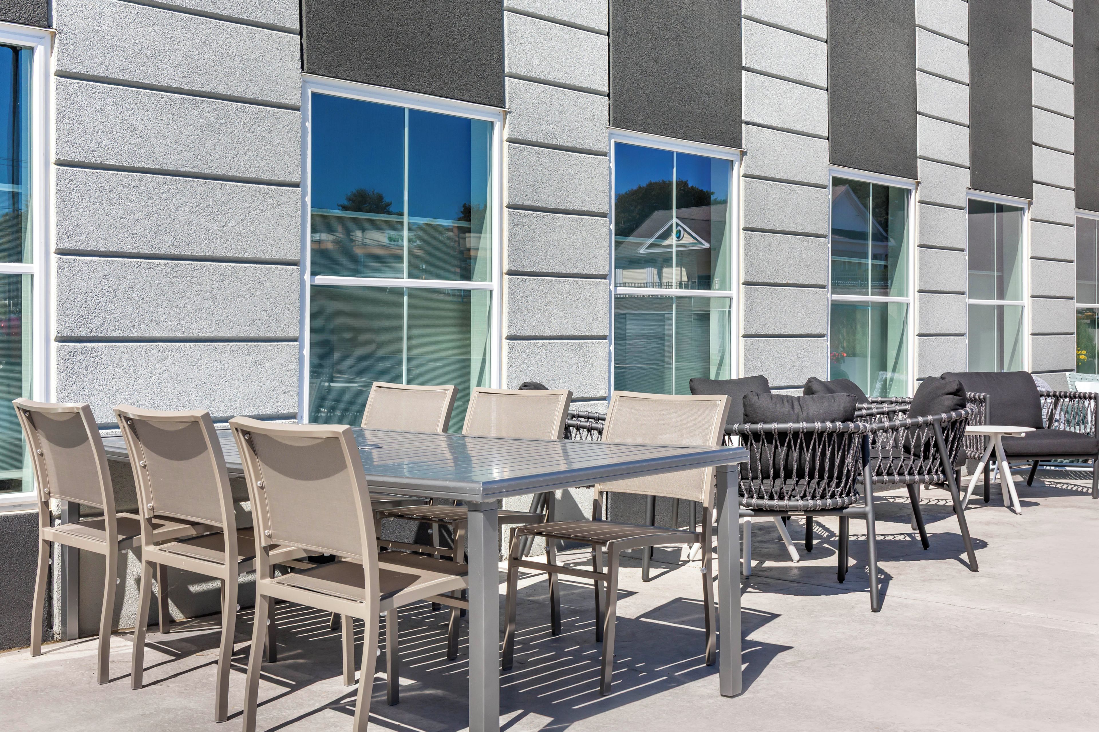Um pátio externo com mesa e cadeiras de vidro, um sofá de vime e vista para um edifício com janelas amplas.
