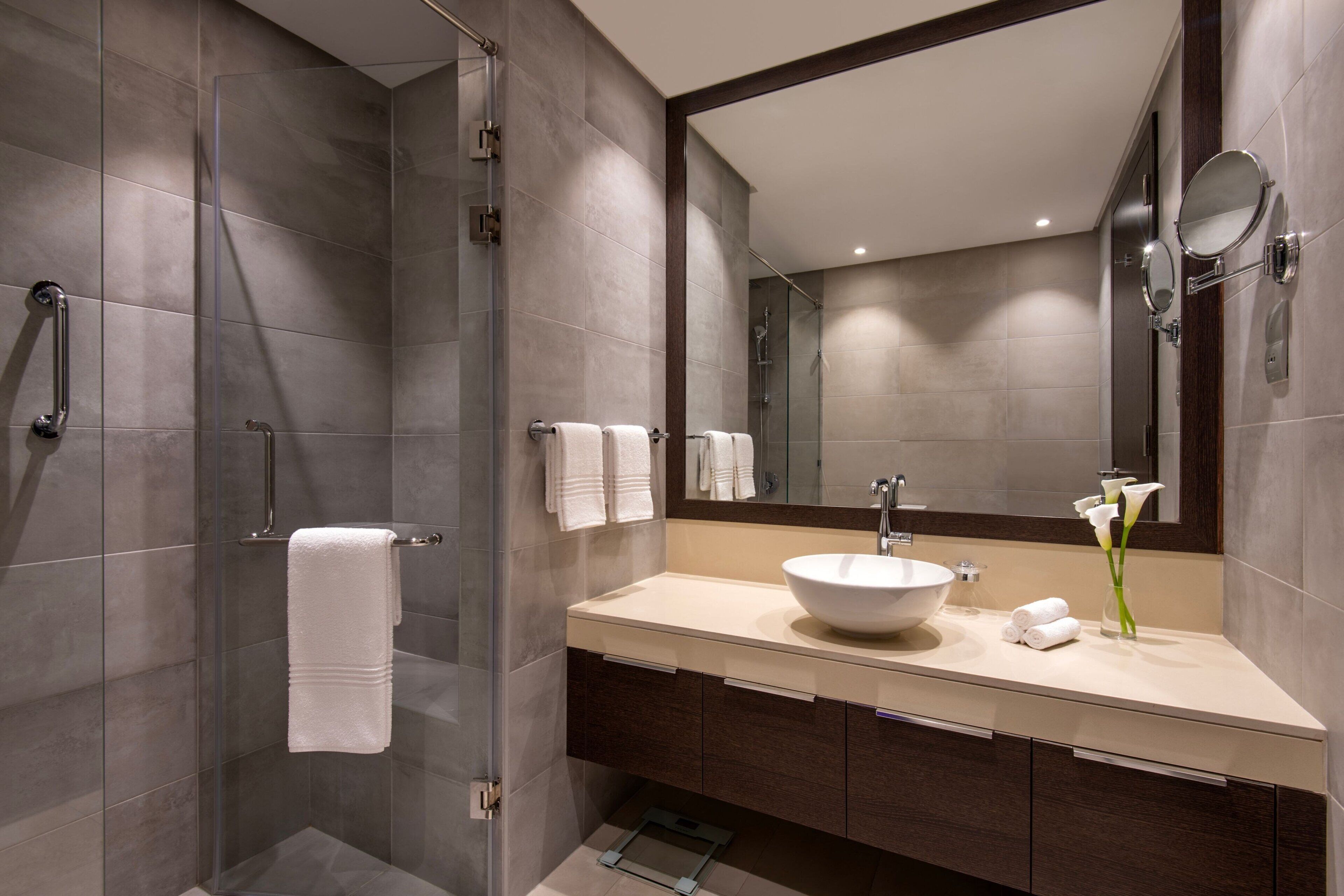 Ein modernes Badezimmer mit einer Glaskabine zur Dusche, einem großen Spiegel, einem Waschbecken und einer Vase mit Blumen.