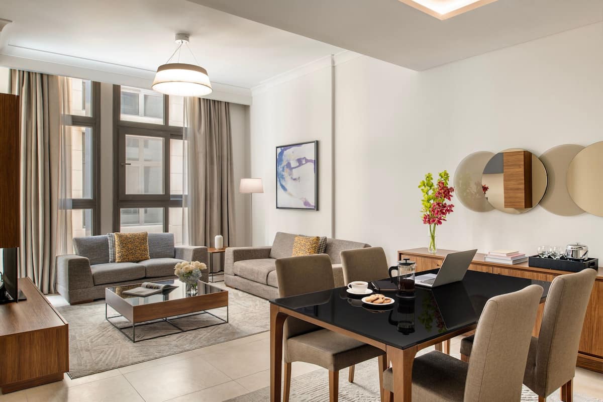 A modern living room with a sofa, coffee table, and dining table. There is a laptop on the table, and a vase with flowers on the side.