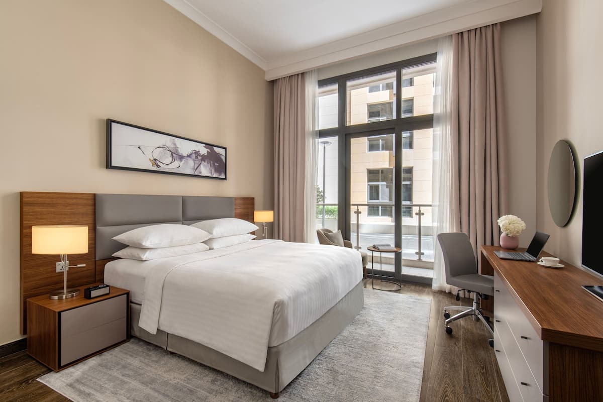 A hotel room with a large bed, a desk with a laptop, a chair, a nightstand with a lamp, and a window with curtains.