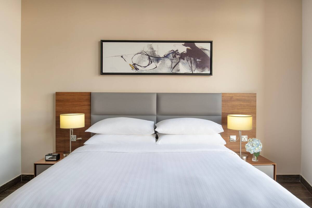 A hotel room with a large bed, two bedside tables with lamps, a framed abstract painting on the wall, and a small vase with flowers on one of the tables.