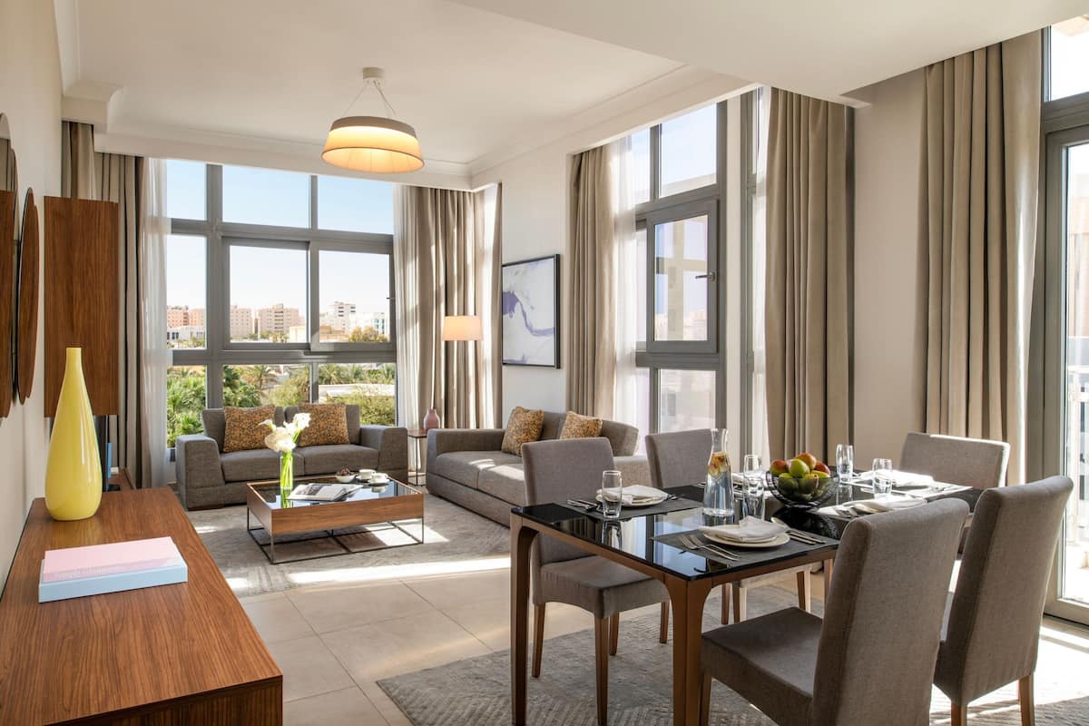 A modern hotel room with a large window, a sofa, a dining table with chairs, and a coffee table.