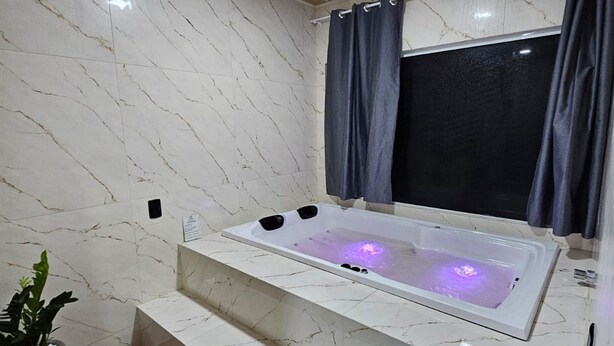 A modern bathroom with a marble floor, a white bathtub with a built-in seat, a small potted plant, and a blue rug. - Refúgio da Montanha - Angelina - SC (Angelina)
