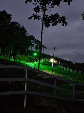 Vista nocturna de una zona rural con un camino iluminado en verde que lleva a una casa.