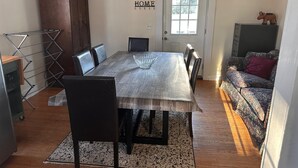 A dining room with a wooden table, chairs, and a decorative bowl. There is a wooden cabinet, a drying rack, and a sofa with a cushion. A door with a window and a sign that reads "HOME" are also visible. - Holmes Residence (Taylorsville)