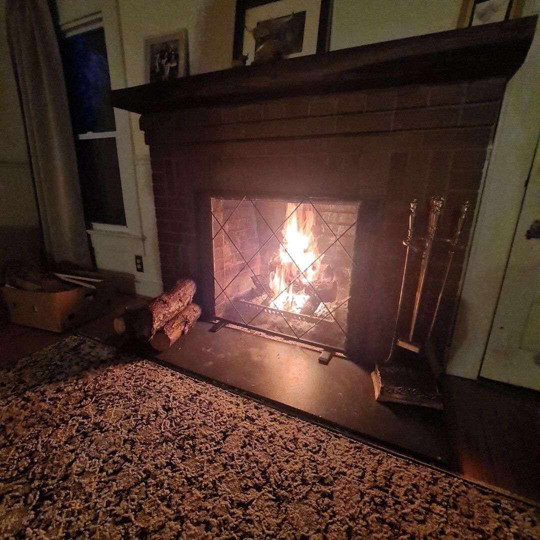 En öppen spis med ved, en matta och ett fönster med gardiner.