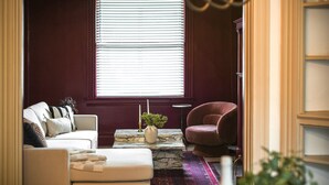 Una sala de estar con un sofá, un sillón rojo, una mesa de centro de mármol y una ventana con persianas.