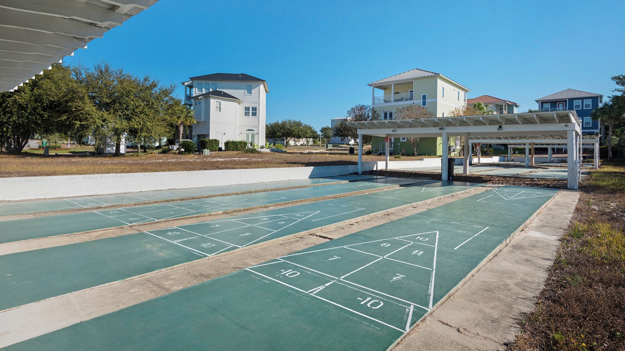 A park with shuffleboard courts, a covered seating area, and residential buildings in the background.
