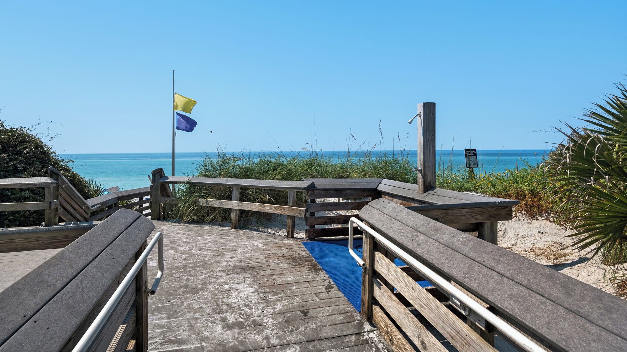 A wooden boardwalk leading to a beach with a flagpole and a sign.