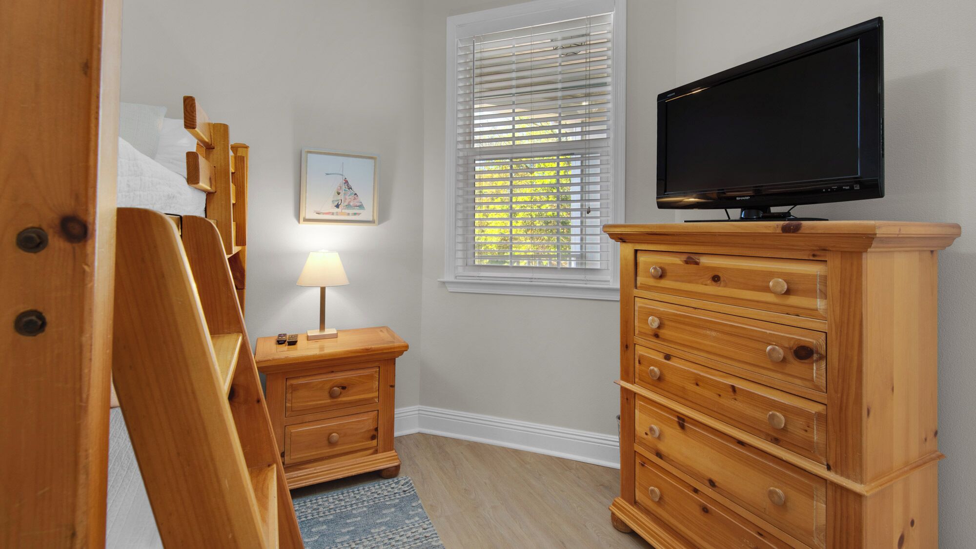A bedroom with a wooden dresser, a television on top, a bedside table with a lamp, and a window with blinds.