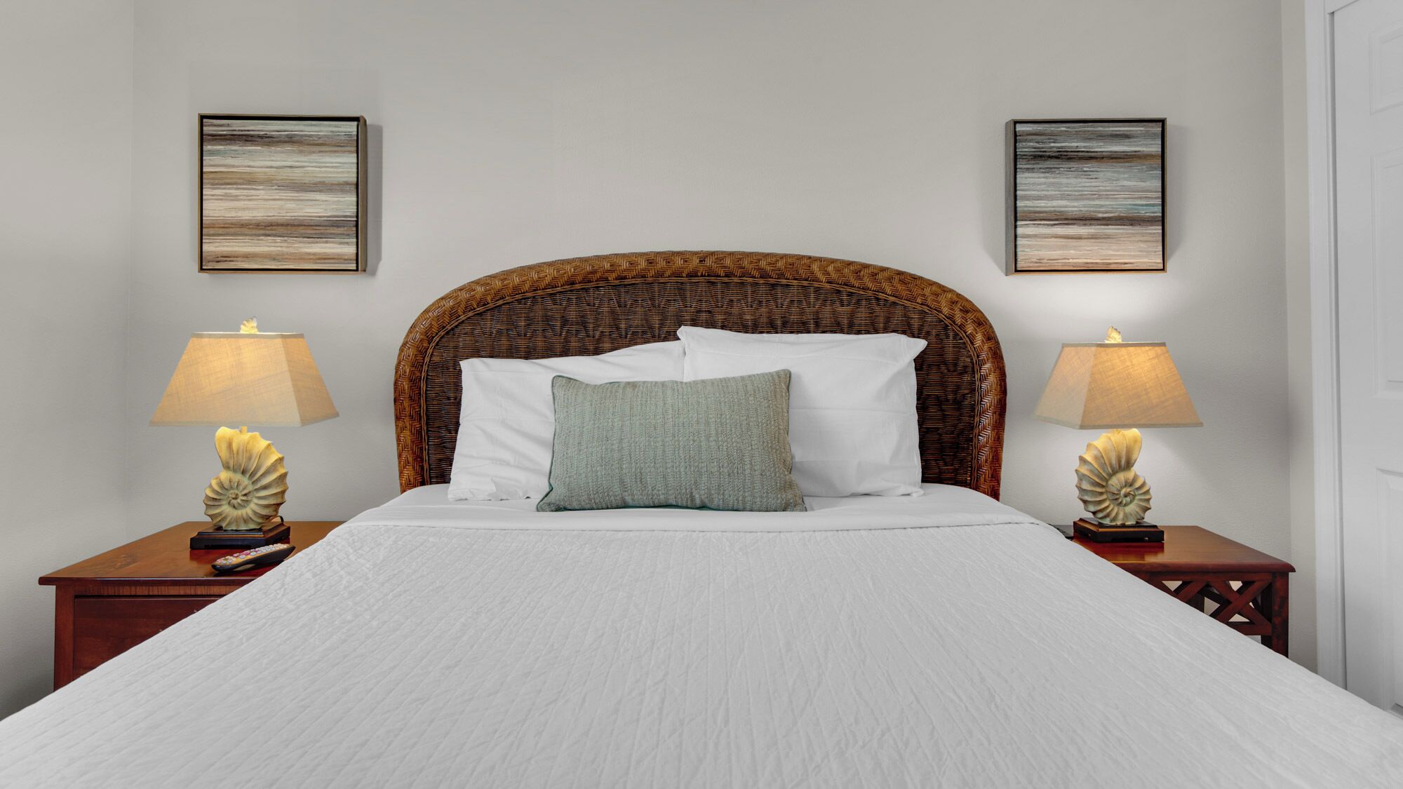 A neatly made bed with a woven headboard, two bedside lamps, and two framed artworks on the walls.