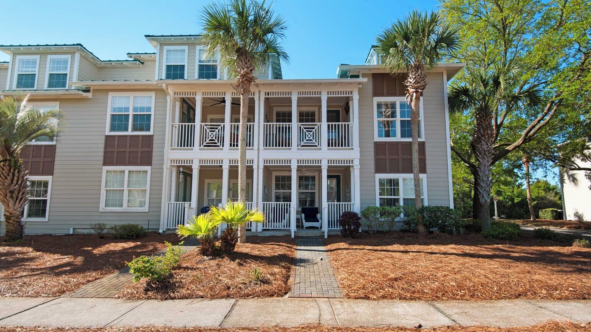 A two-story building with a central balcony, a covered porch, and a landscaped front yard with palm trees and shrubs.
