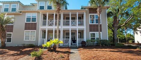 A two-story building with a central balcony, a covered porch, and a landscaped front yard with palm trees and shrubs.